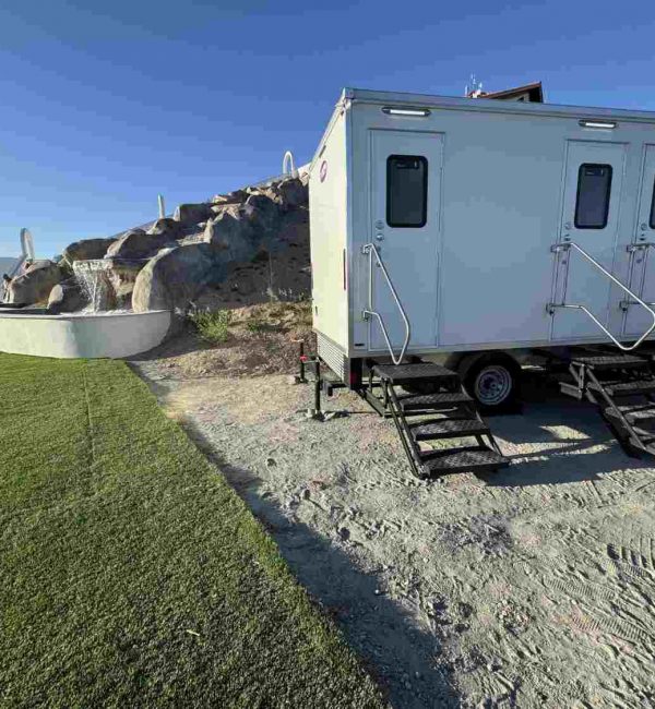 White three-stall restroom trailer with stairs and handrails set up beside a lawn and decorative rock waterfall.