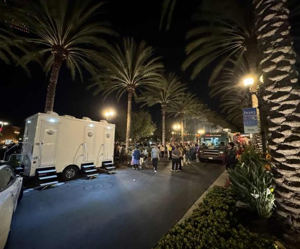 Crowded nighttime outdoor event lined with illuminated palm trees, food trucks, and a mobile restroom trailer.