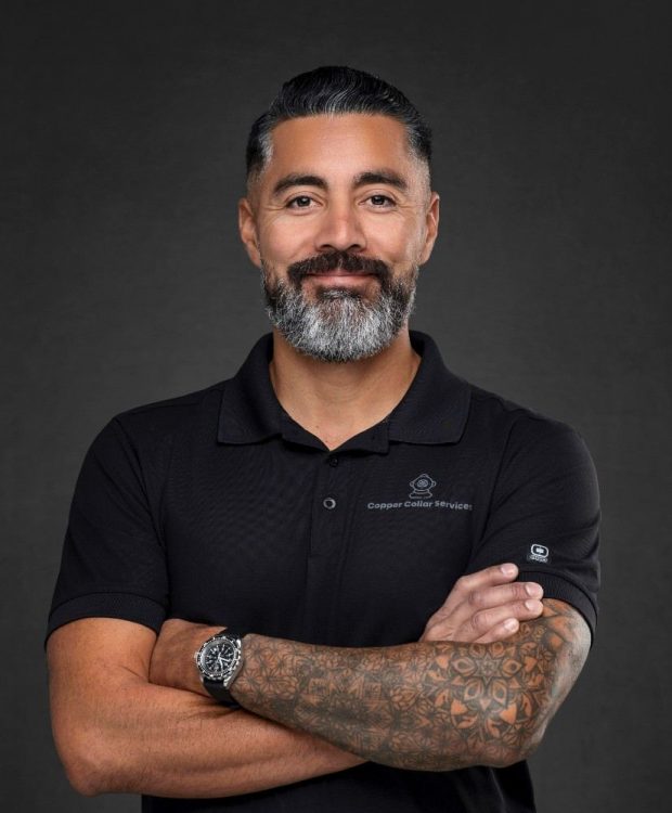 Professional portrait of a bearded Copper Collar Services team member wearing a black polo shirt with his arms crossed against a dark background.